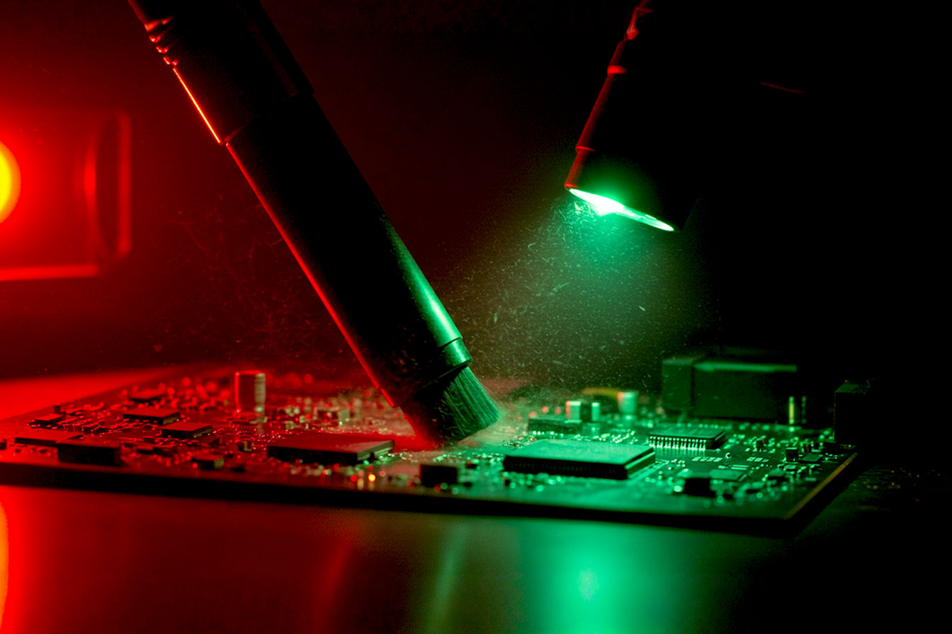 A close-up professional photo of an ESD-safe vacuum with specialized attachments gently cleaning a computer motherboard. The image shows the precision nozzle hovering slightly above the surface while dust particles are being pulled away from components, all on a clean anti-static mat with proper grounding equipment visible.