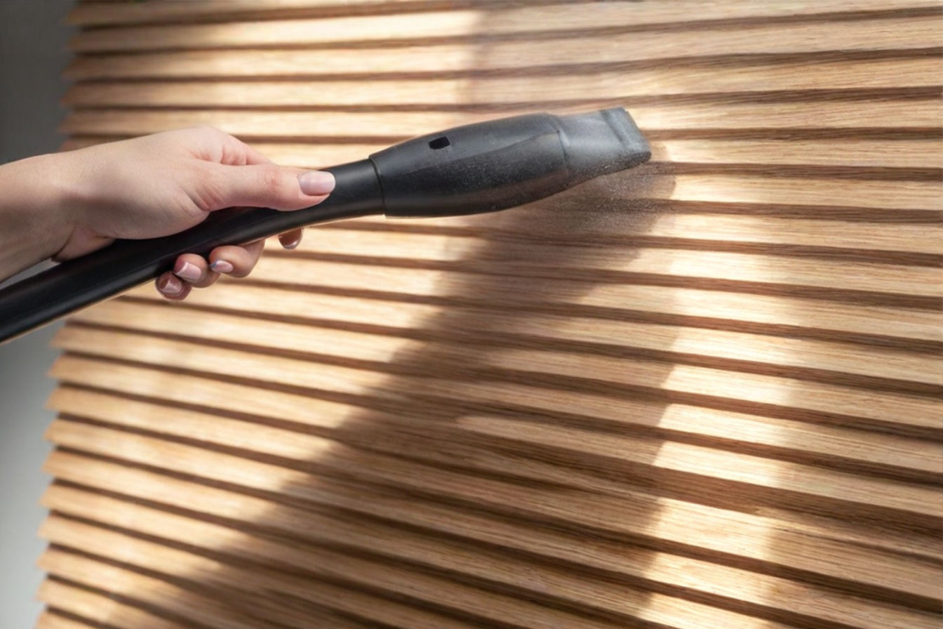 A close-up professional photograph of a hand using a specialized vacuum attachment to clean horizontal wooden blinds. The image shows dust being effectively removed by the brush attachment in natural lighting, with visible dust particles being captured by the vacuum. The composition shows both the cleaning tool and a section of clean versus dusty blinds for contrast.